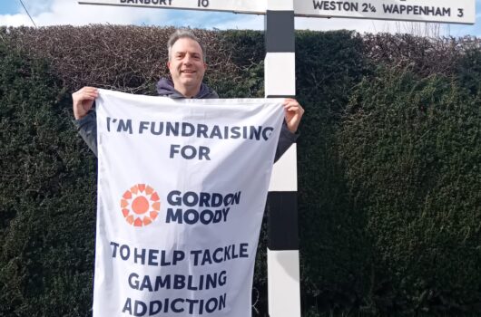 Dave Hollingsworth standing in front of a finger post road sign whilst holding his Gordon Moody fundraising flag showing how to donate to his challenge.